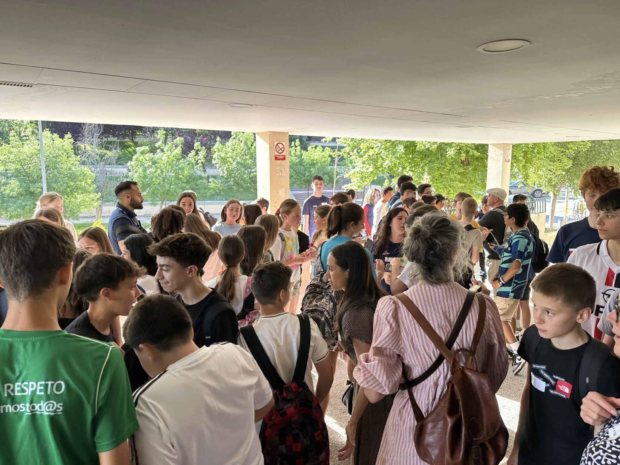 A large group of people, mainly teenagers, gather under a covered area in a park-like setting, engaging in conversation.