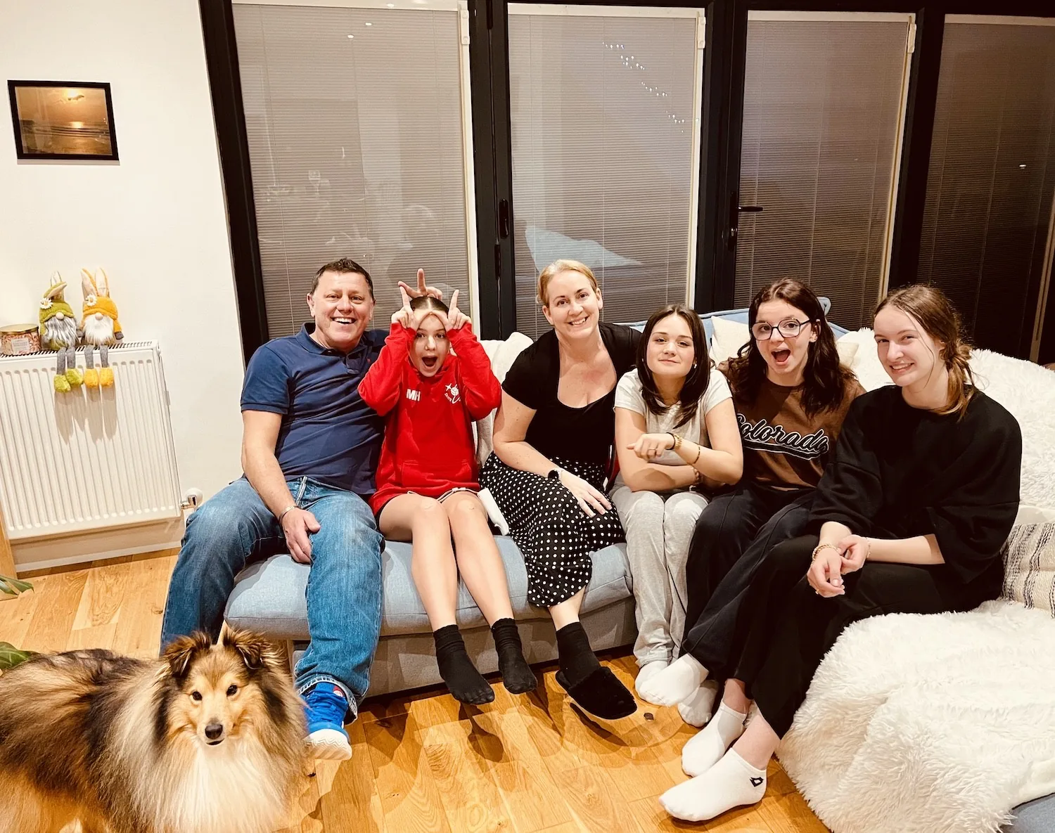 Family smiling together on a couch with a dog in the foreground; indoor setting with decorative gnomes on a radiator.